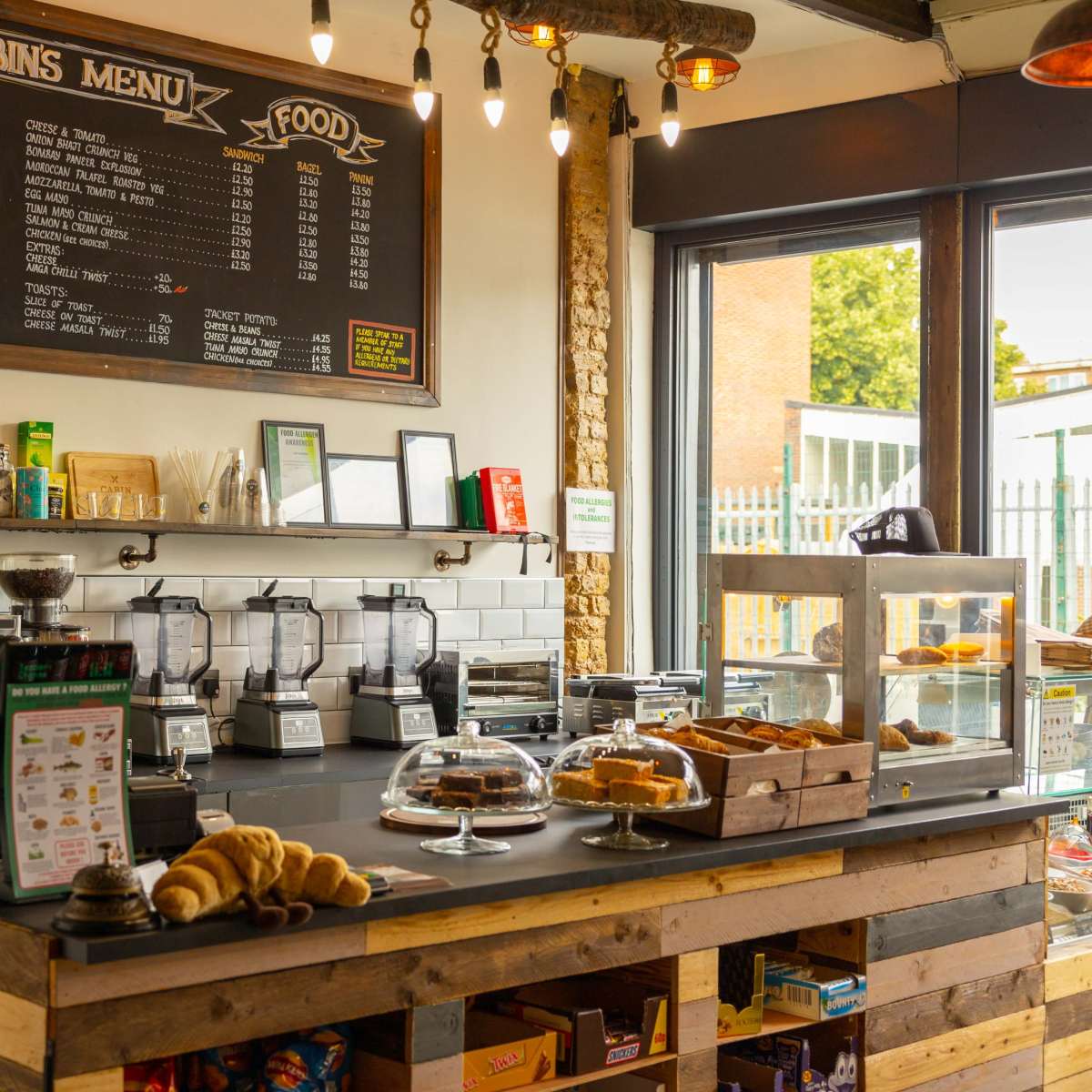 A cozy café interior featuring a menu board, a coffee machine, glass display cases with baked goods, and a rustic wooden counter.