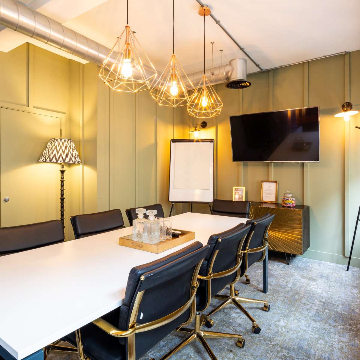 A conference room features a rectangular white table surrounded by black chairs, with pendant lights overhead and a large wall-mounted screen.