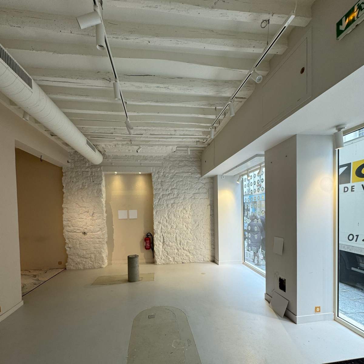 An empty commercial space with white walls, exposed ceiling beams, and large windows facing the street.