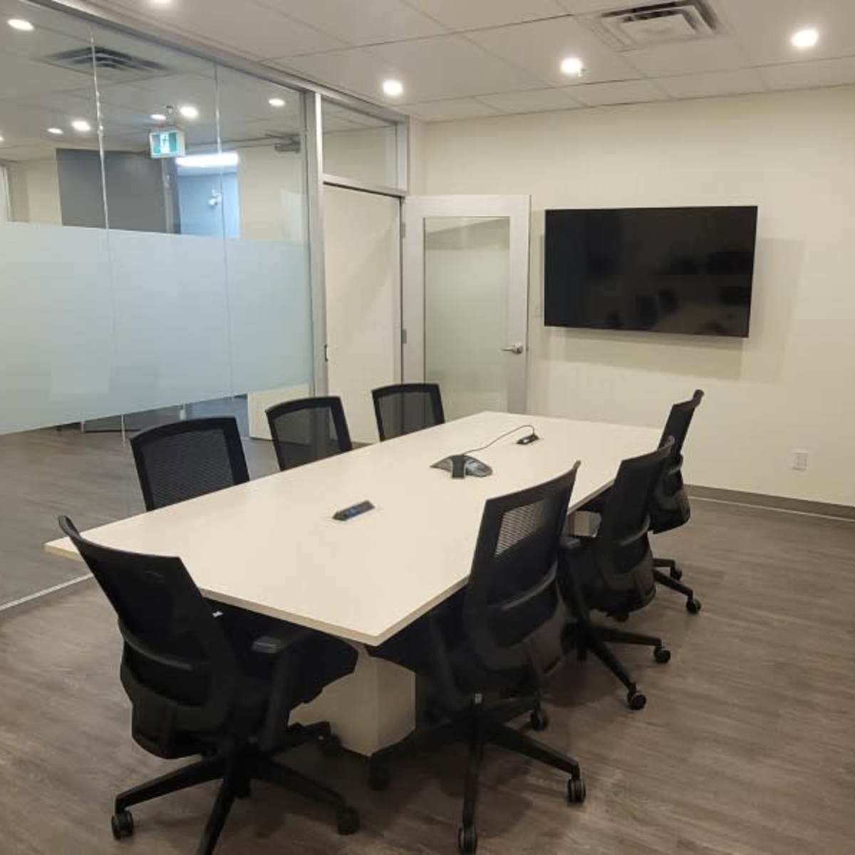 A modern conference room with a long rectangular table surrounded by six black chairs, two wall-mounted screens, and large glass windows.