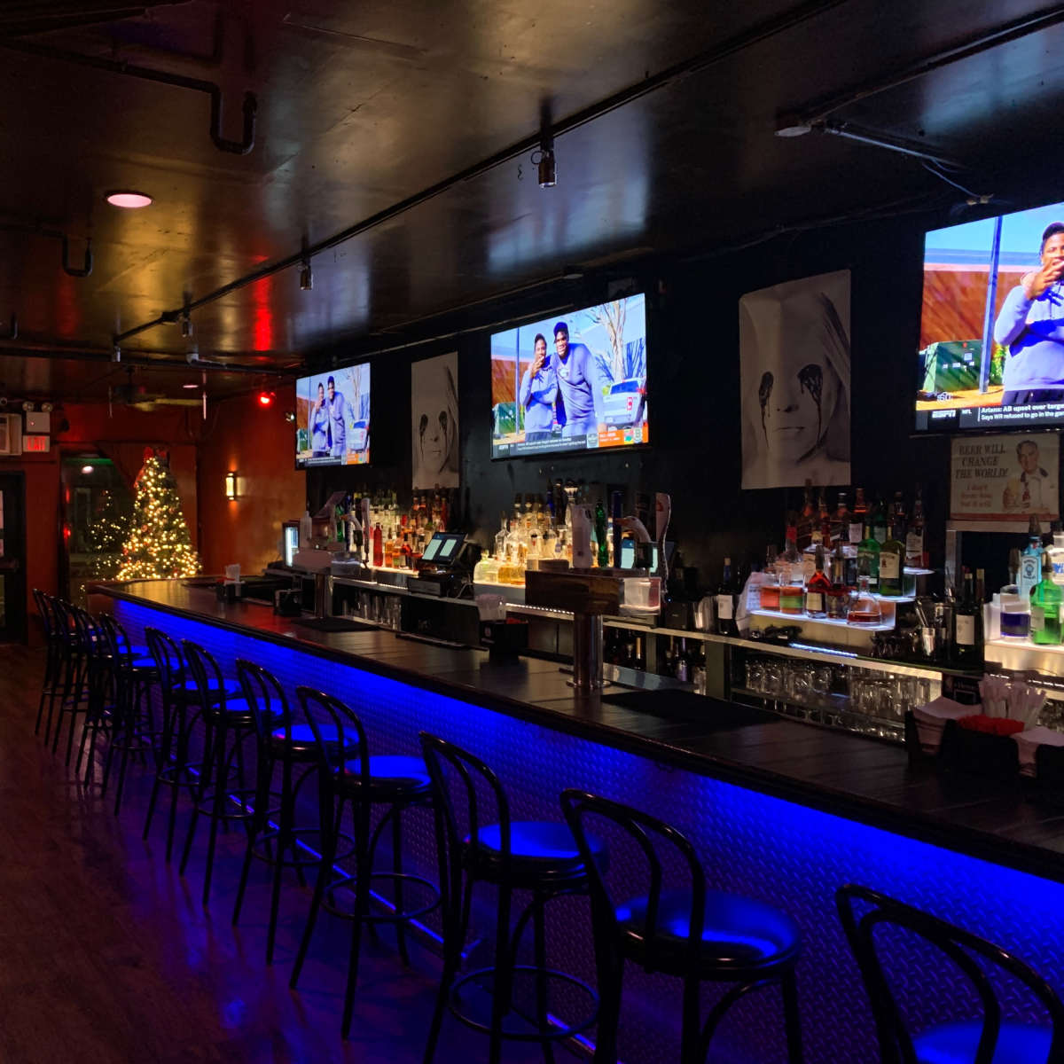 A dimly lit bar with multiple televisions displaying a program, a decorated Christmas tree, and a long, illuminated bar counter lined with chairs.