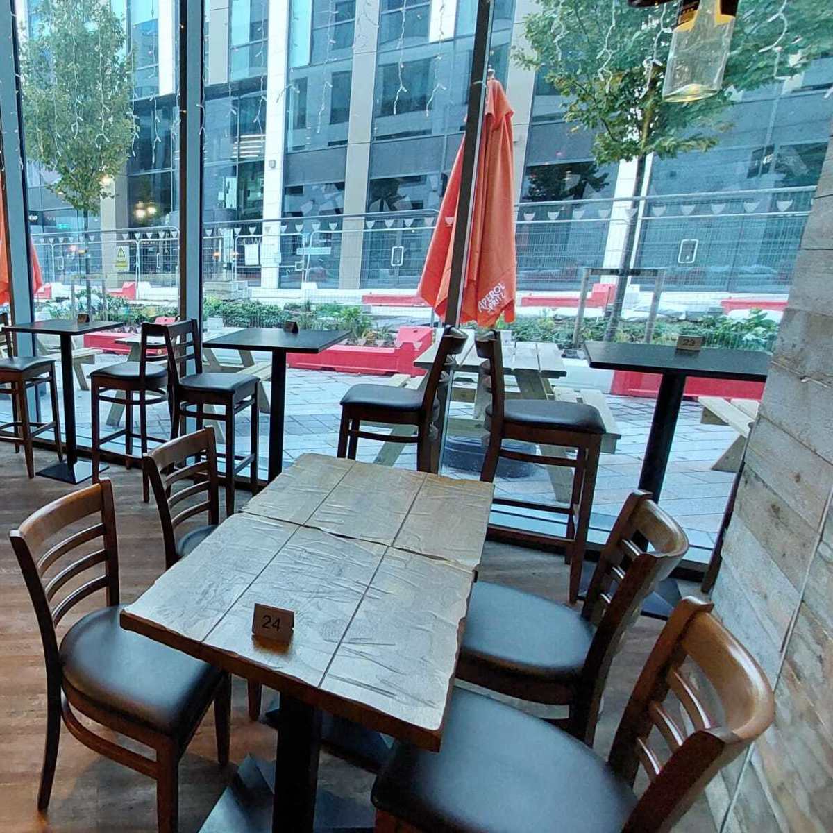 A modern restaurant interior with wooden tables and chairs arranged near large windows overlooking a city street.