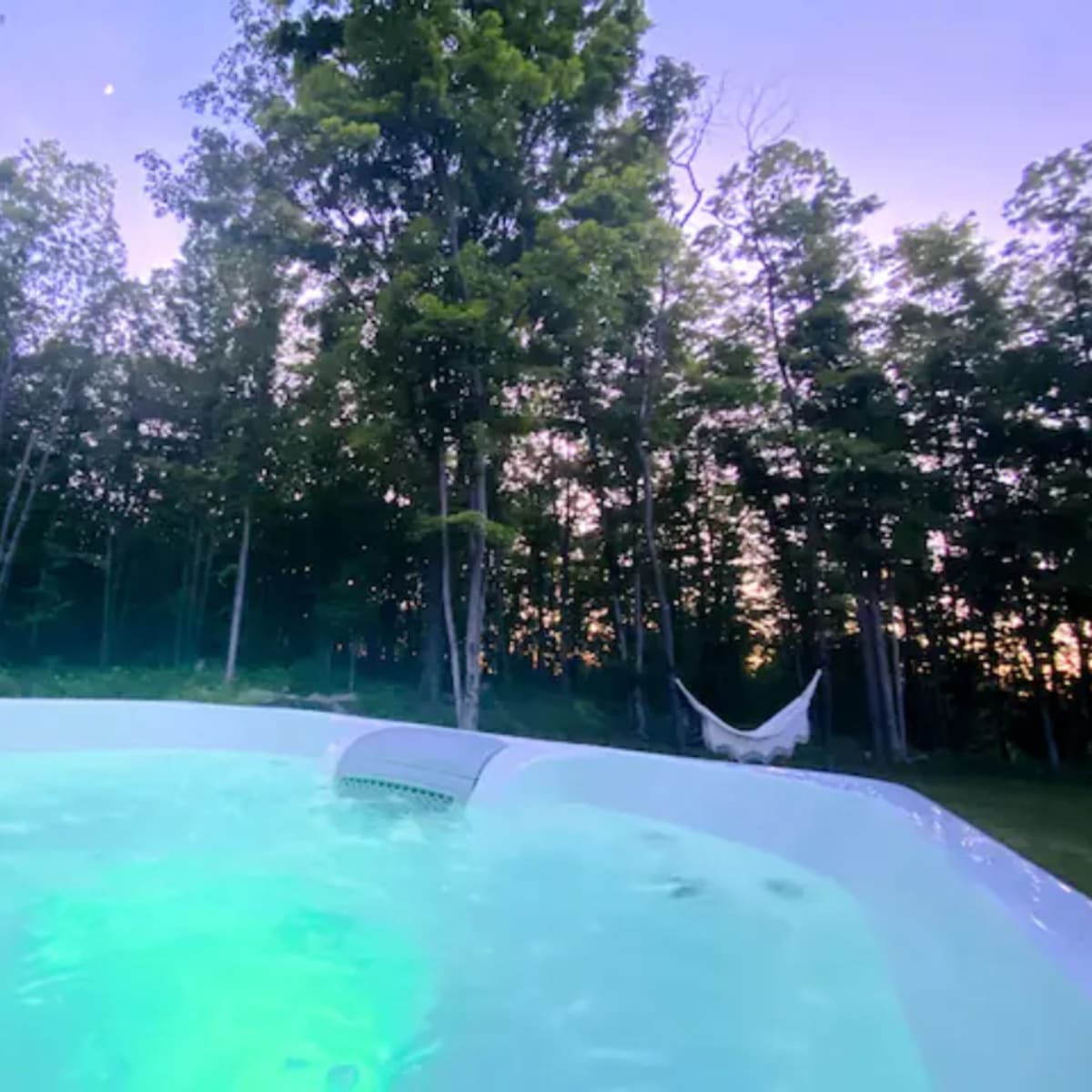 A swimming pool illuminated with green light, surrounded by trees and a hammock in a forested area during twilight.