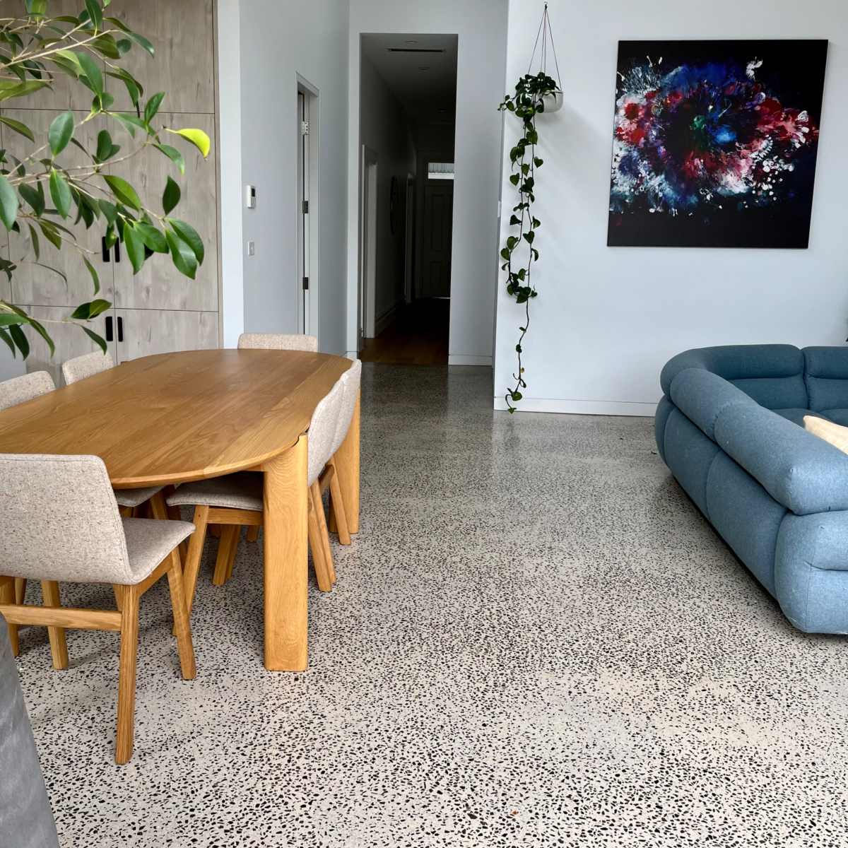 A modern living space with a dining area featuring a wooden table and four chairs, a blue sectional sofa, and a wall adorned with a colorful abstract painting.