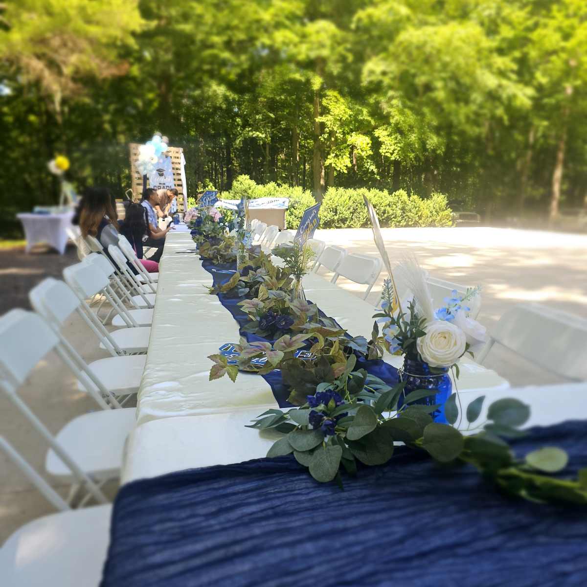 A long, decorated table with a blue runner and floral arrangements is set up for an outdoor event in a green, wooded area.