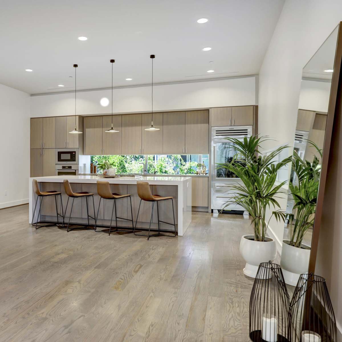 A modern kitchen features a central island with three pendant lights, sleek cabinetry, and large windows overlooking greenery.