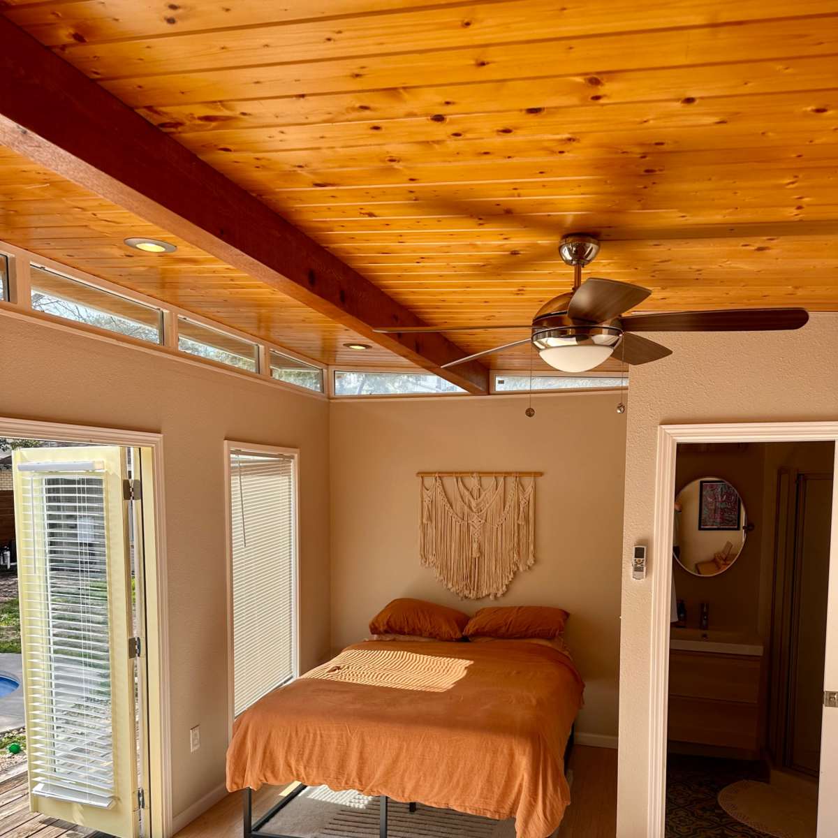 A small bedroom features a bed with an orange blanket, wooden ceiling beams, and large windows allowing natural light.