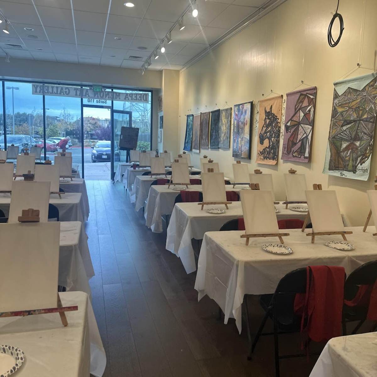A spacious art studio with tables covered in white cloths, each set up with blank canvases and palettes, surrounded by colorful artwork displayed on the walls.