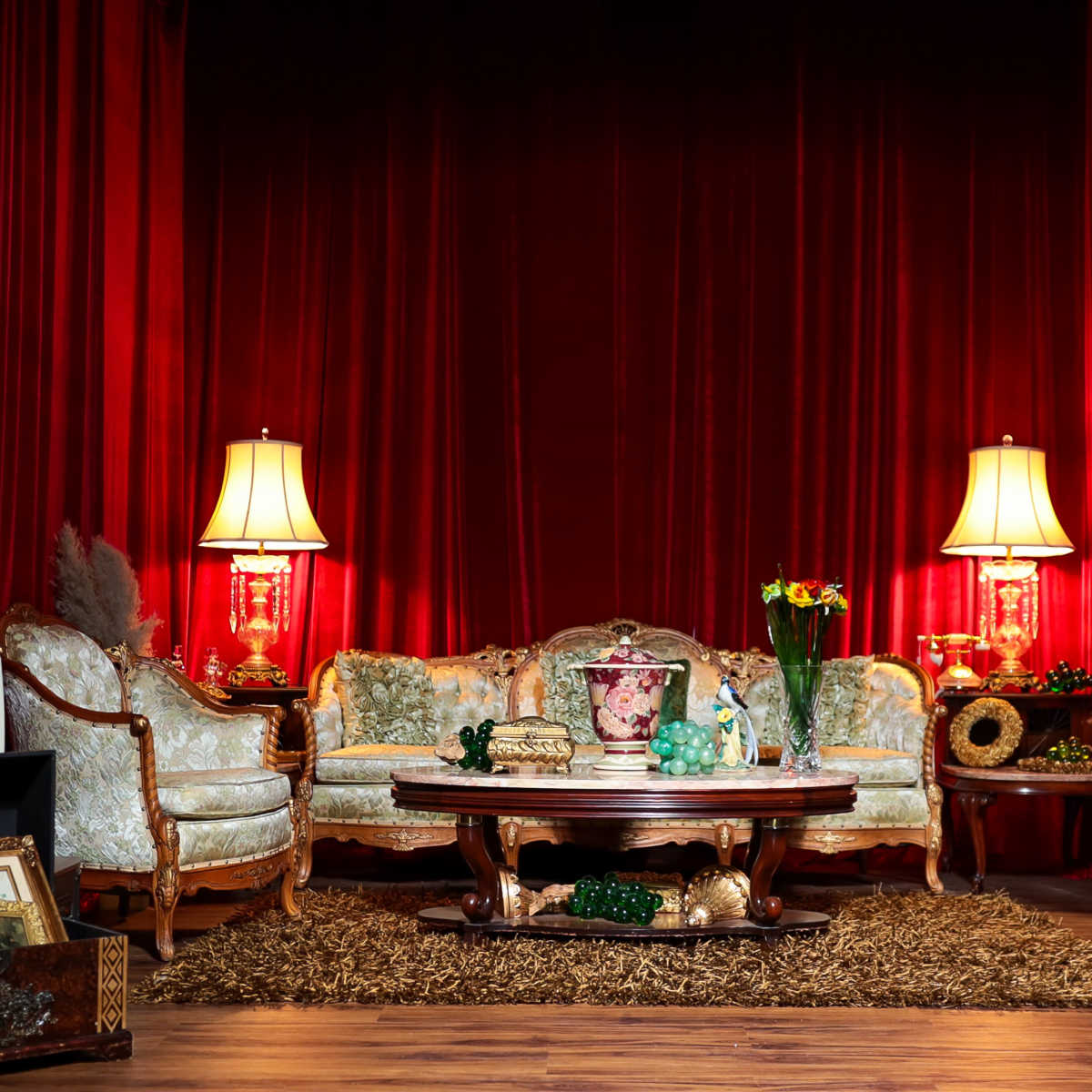 A vintage living room setting features a floral-patterned couch and armchairs, a wooden coffee table, two lamps, and various decorative objects, all set against a backdrop of red curtains.