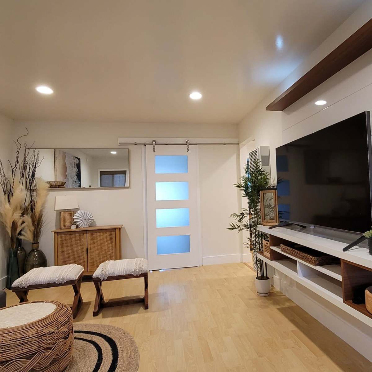 The living room features a contemporary design with a wall-mounted television, a sliding glass door, and natural decor elements like wicker baskets and potted plants.
