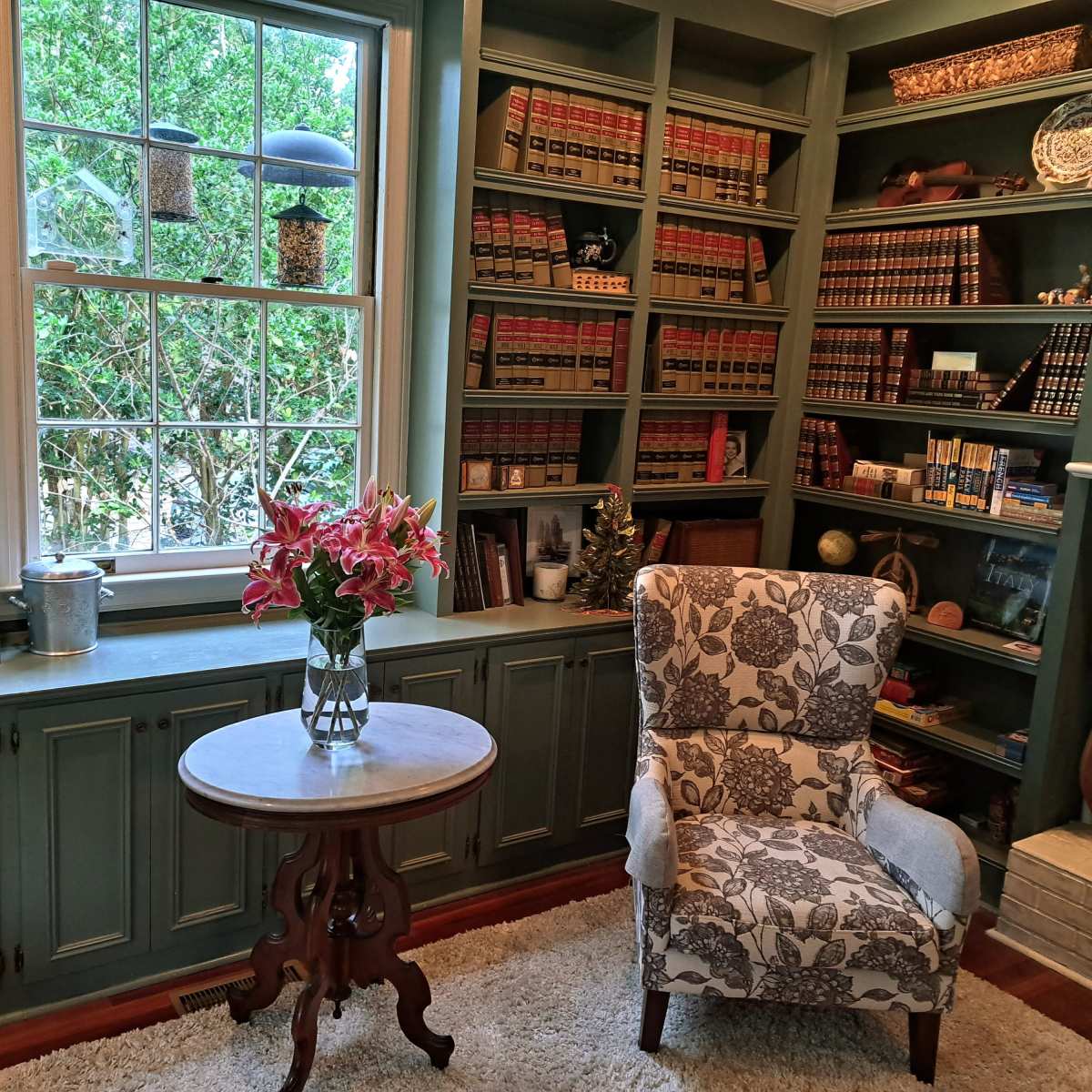 A cozy reading nook features a patterned armchair beside a round table, with bookshelves filled with law books and a window letting in natural light.