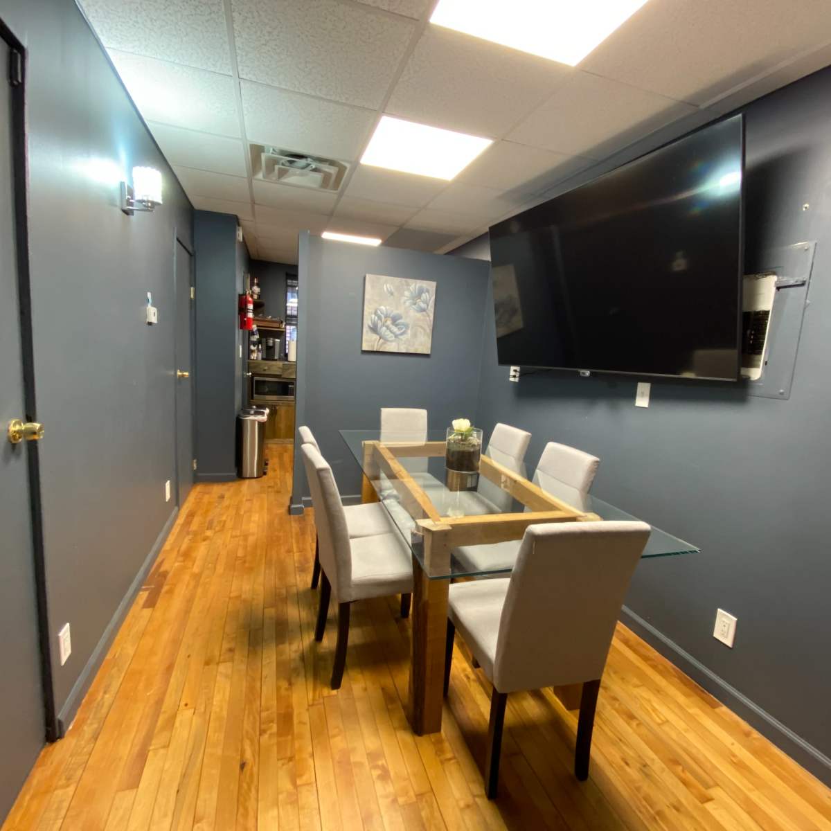 A modern conference room with a rectangular glass table surrounded by six upholstered chairs, a large wall-mounted TV, and gray-painted walls.