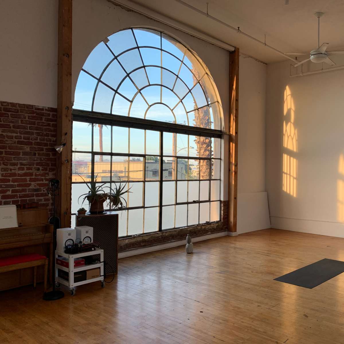 A spacious room with a large arched window that casts shadows on the wooden floor, featuring a yoga mat and a few plants.