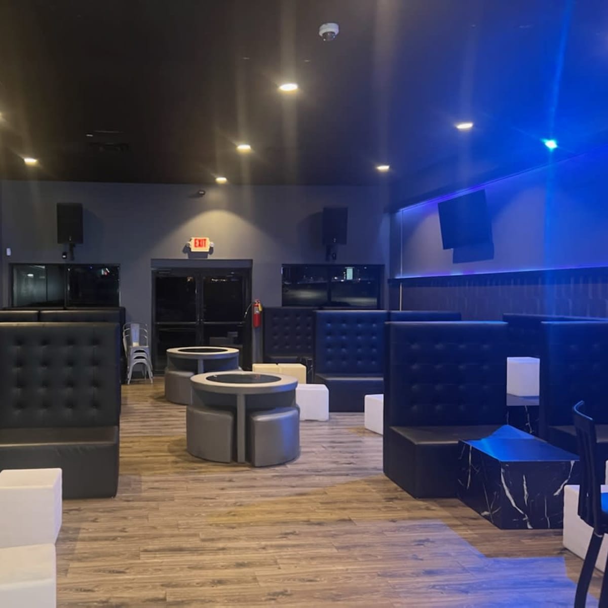 A dimly lit lounge area featuring black upholstered seating, round tables, and a wooden floor.