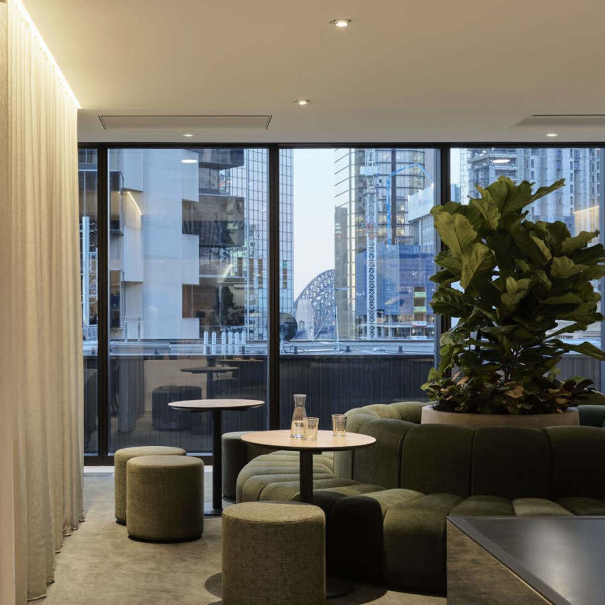 A modern lounge area features green seating, small tables, and large windows with city views.