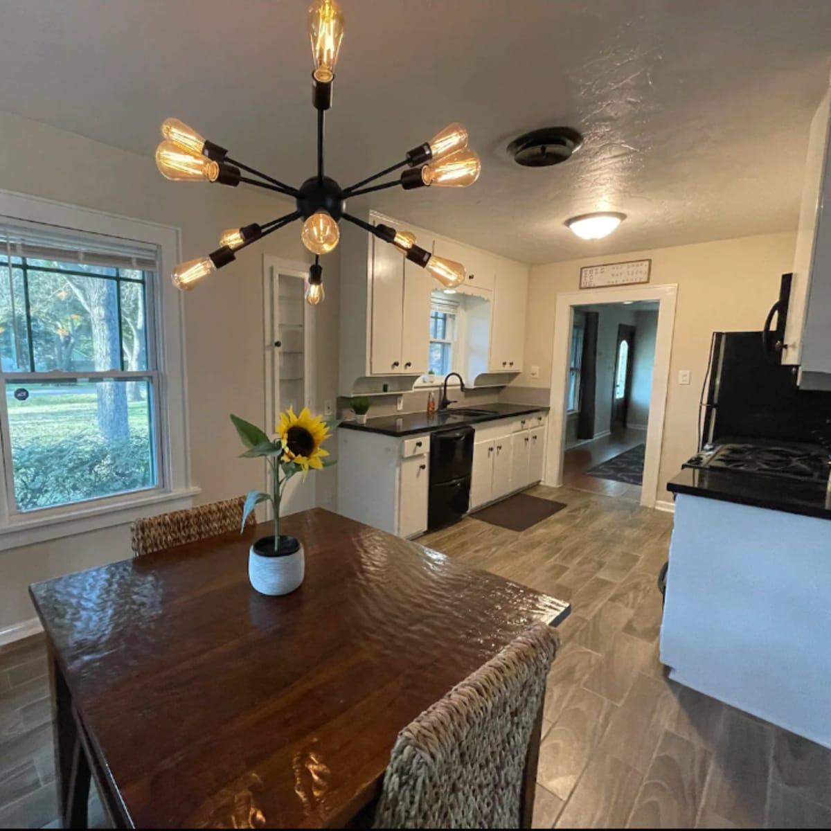 A kitchen features a wooden dining table with a sunflower centerpiece, a black countertop, and a modern light fixture overhead.
