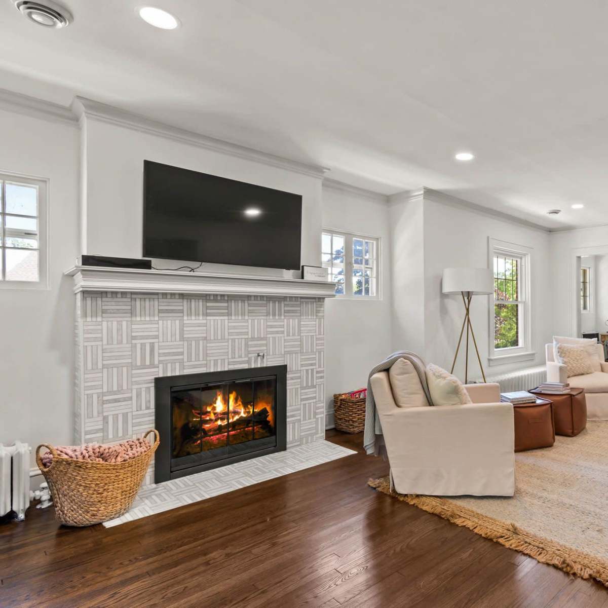 A living room featuring a fireplace with a television mounted above it, two armchairs, a coffee table, and a woven rug on hardwood flooring.