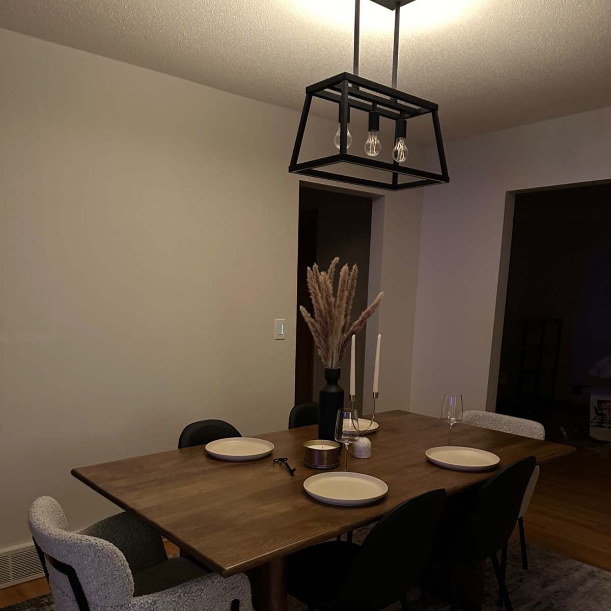 A wooden dining table set for four with plates, candles, and a vase of dried flowers is surrounded by black chairs in a softly lit room.