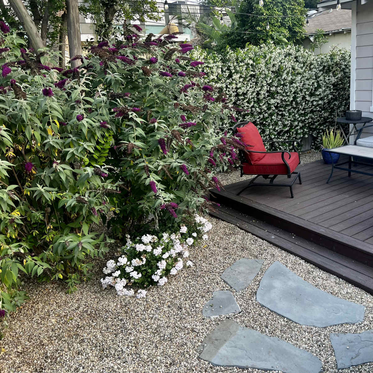 A small outdoor seating area features a wooden deck with two red chairs and a sofa, surrounded by flowering bushes and gravel pathways.