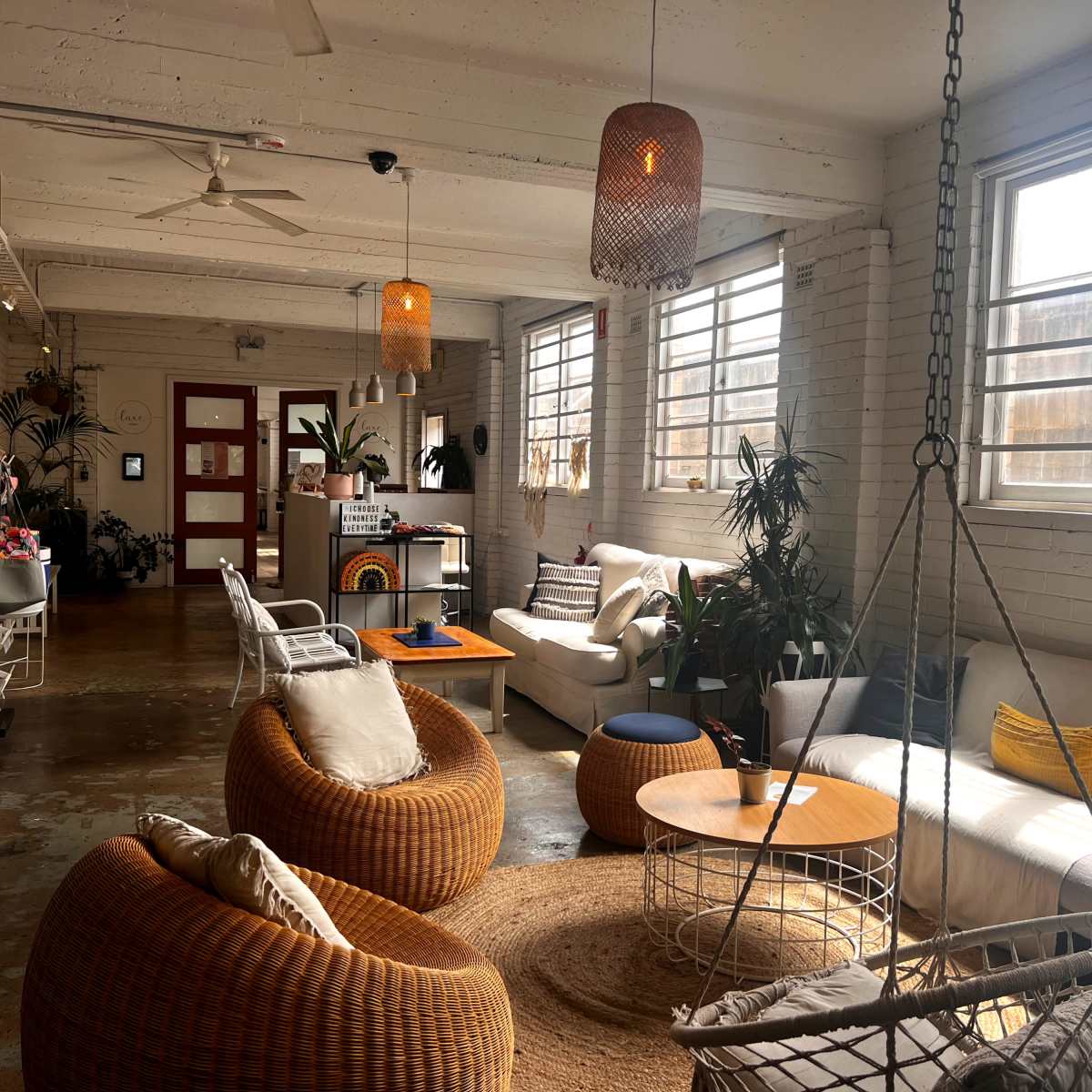 A spacious, well-lit lounge area featuring a variety of seating options, including woven chairs and sofas, along with potted plants and large windows.