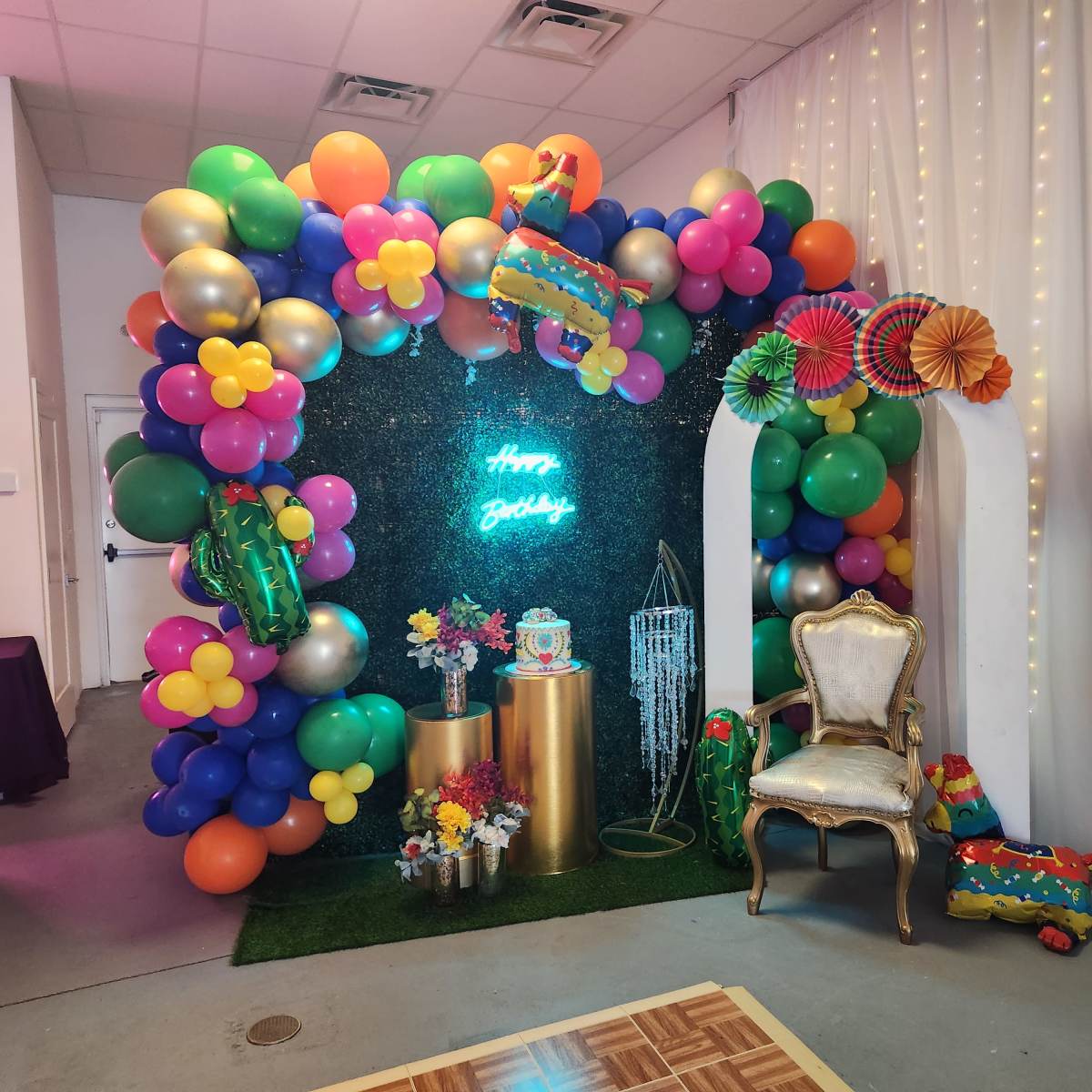A festive birthday backdrop featuring colorful balloons, a decorated table with flowers, and a chair surrounded by party decorations.