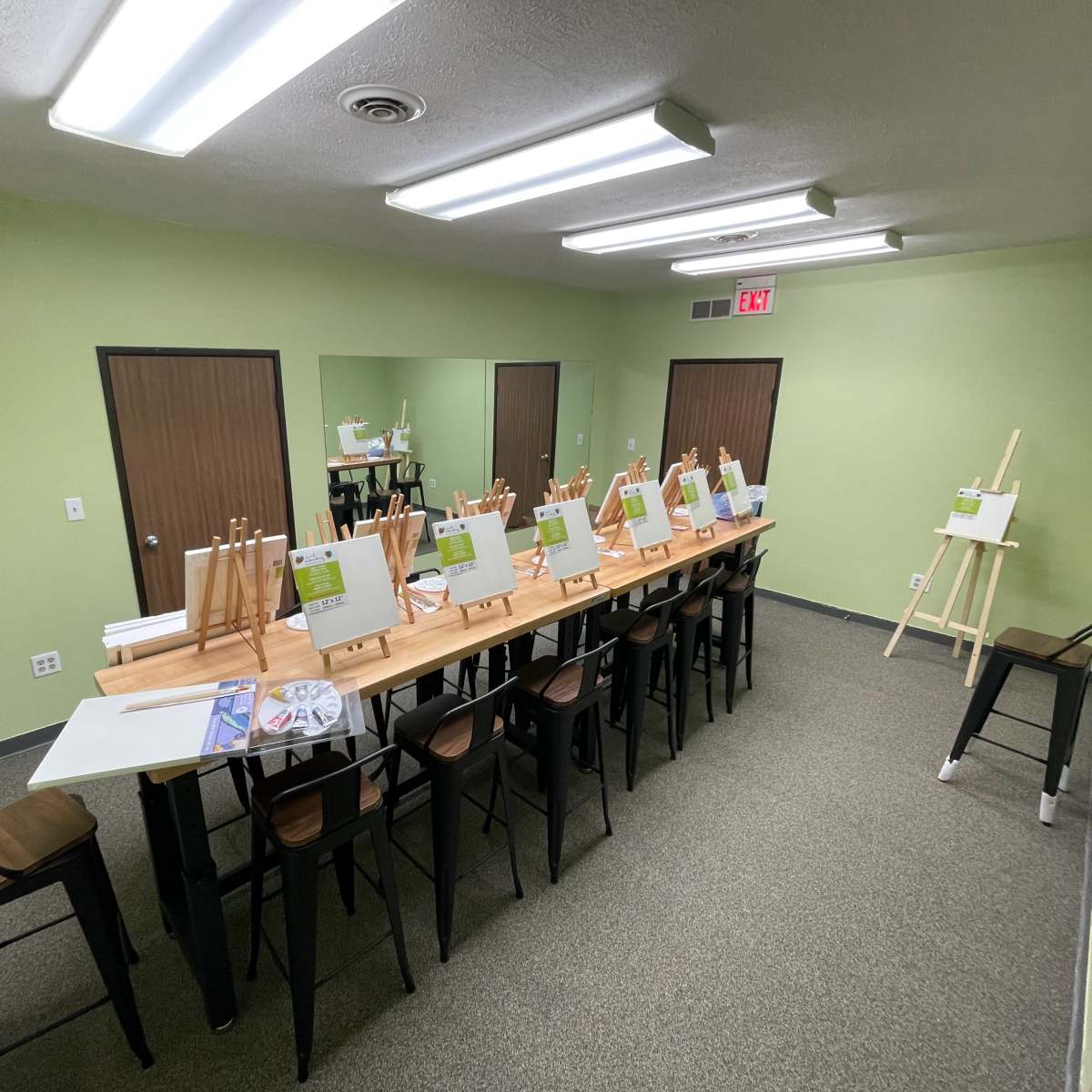 A spacious studio is set up with a long table lined with easels and blank canvases, surrounded by chairs and mirrors on the walls.