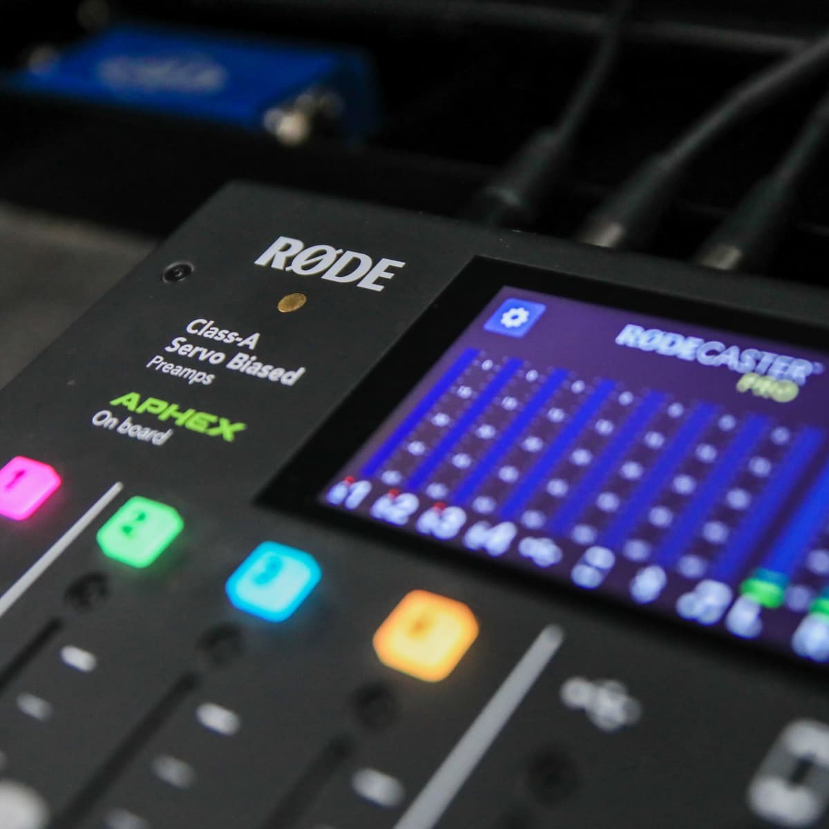 A close-up of a Rode audio mixing console with a digital display, featuring various colored buttons and audio sliders.