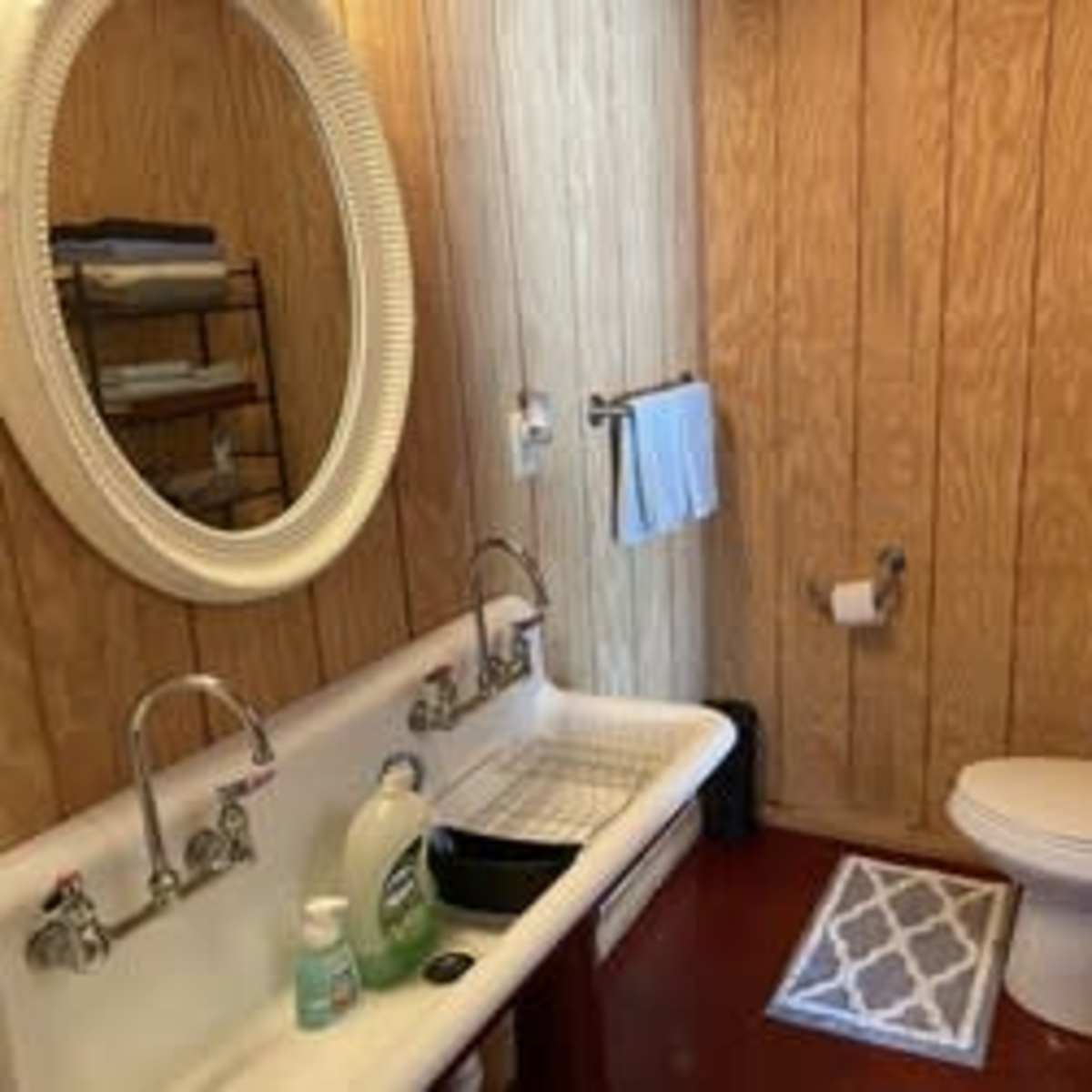 The bathroom features a vintage sink with a countertop, a toilet, a shelf with neatly arranged towels, and wooden paneling on the walls.