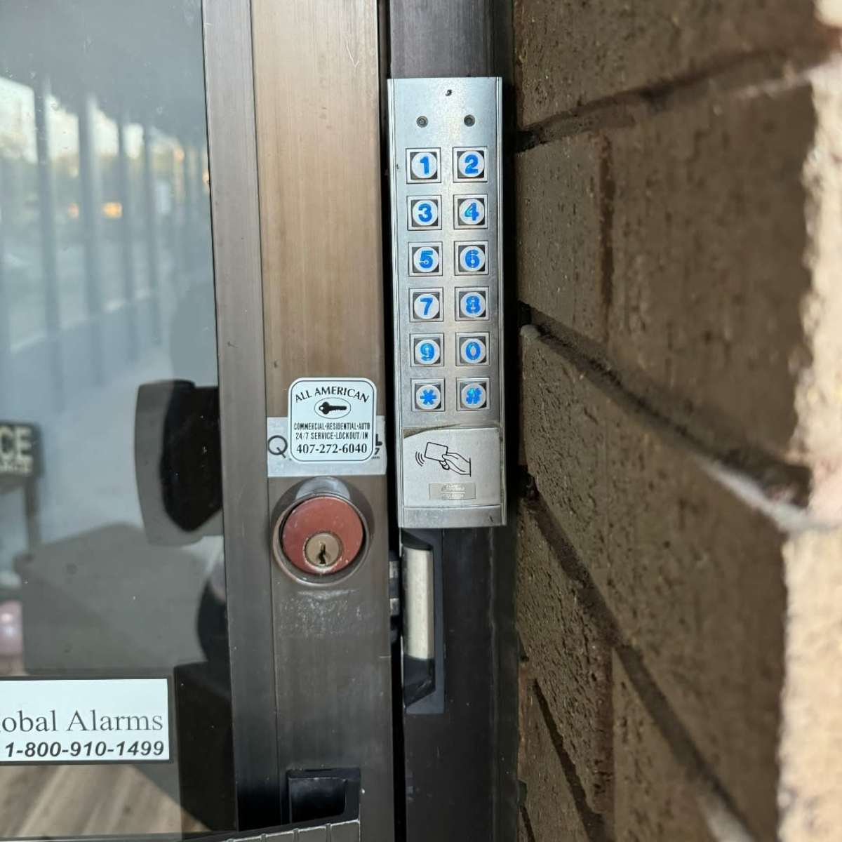 A keypad mounted on a doorframe, featuring numeric buttons and a card reader, alongside an alarm system sticker.