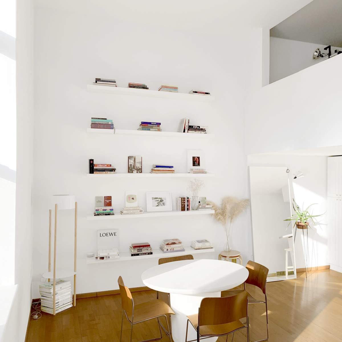 A bright, minimalistic living space with a circular table surrounded by wooden chairs, a tall shelf displaying books, and a kitchenette in the background.
