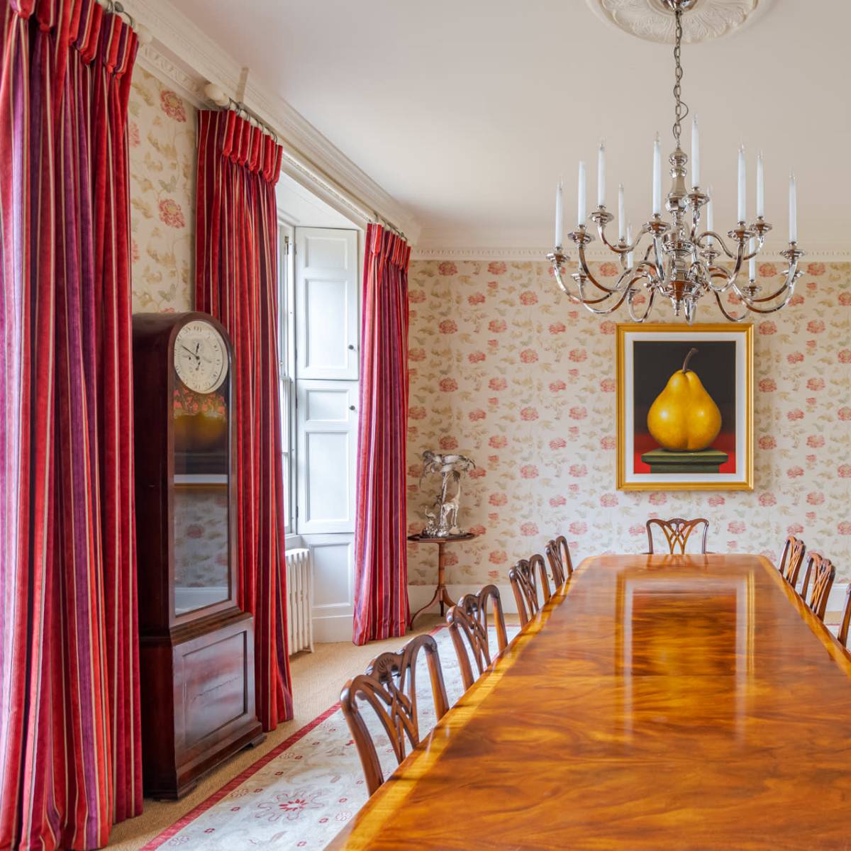 A long wooden dining table is set in an elegantly decorated room with floral wallpaper, framed artwork, and a chandelier hanging above.