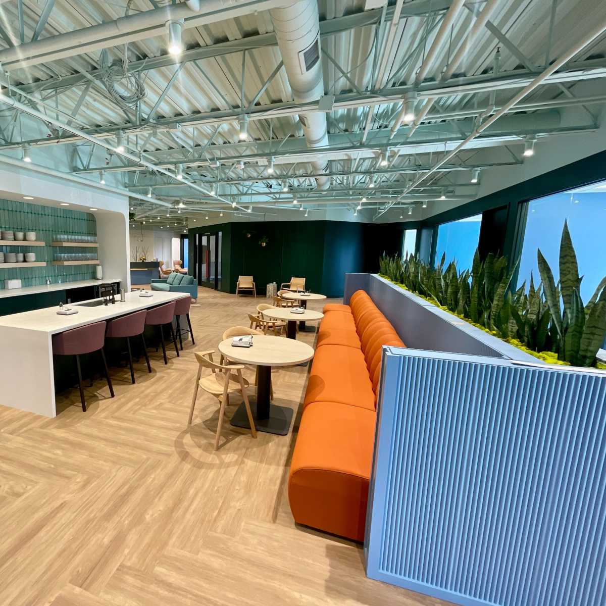 A modern office lounge area with a long orange couch, several round tables, and a kitchen counter lined with colorful dishware.