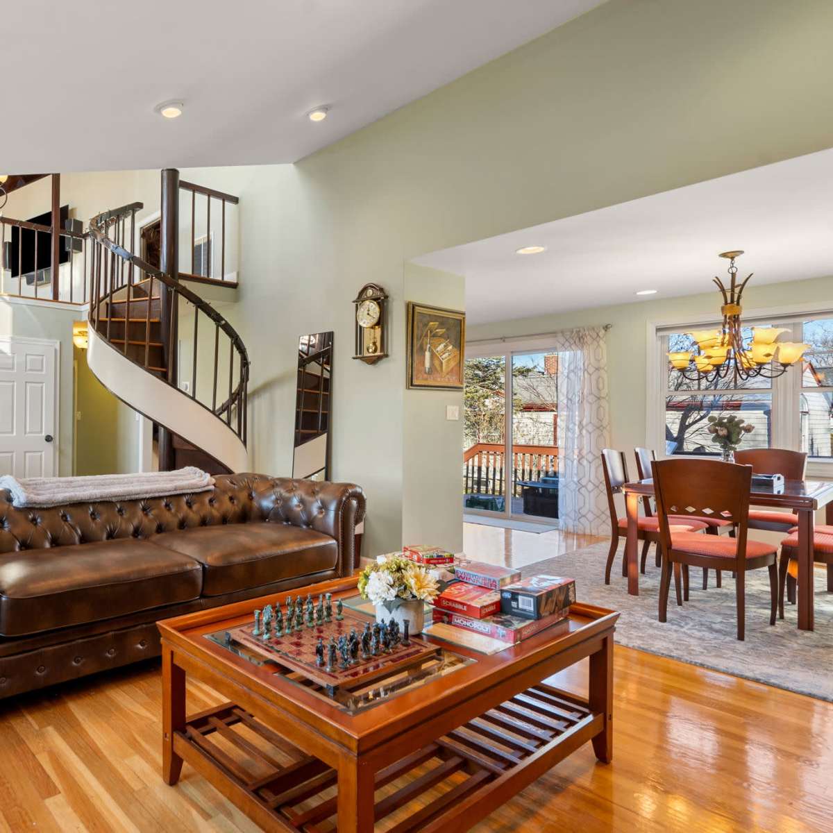 A living room with a leather chesterfield sofa, a wooden coffee table with a chess set on it, and a spiral staircase leading to the upper floor.