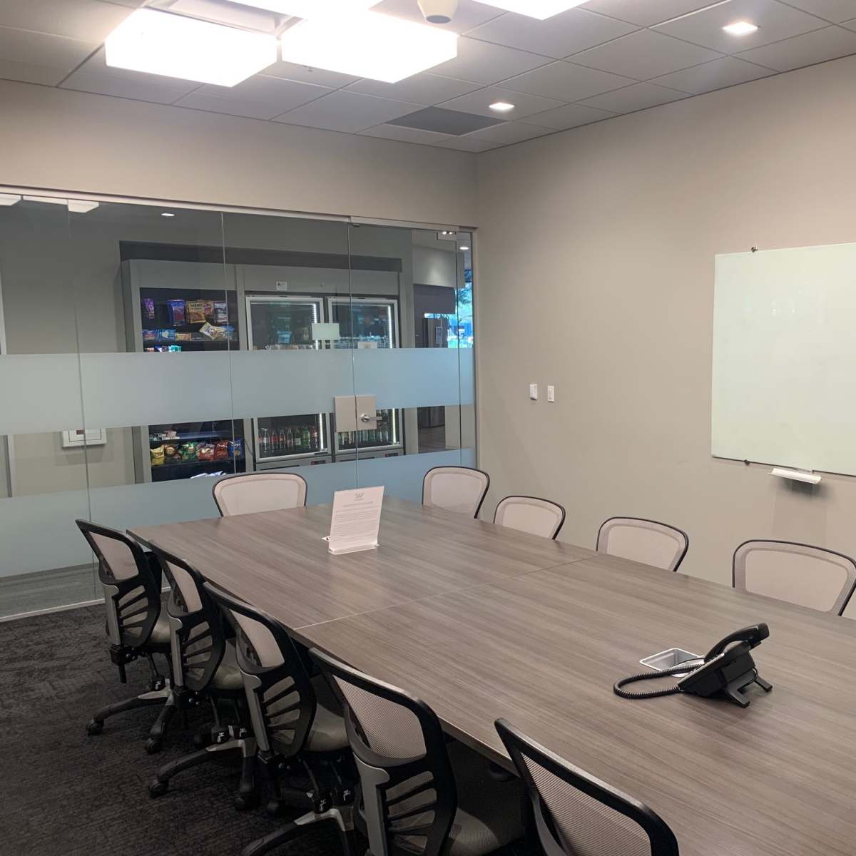 A conference room features a long wooden table surrounded by chairs, with a whiteboard on one wall and a glass partition showcasing a beverage fridge.