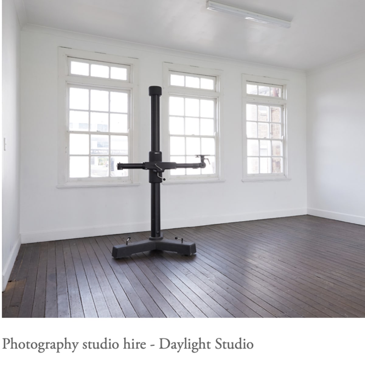 A minimally furnished photography studio with large windows and a central mounting stand on a wooden floor.
