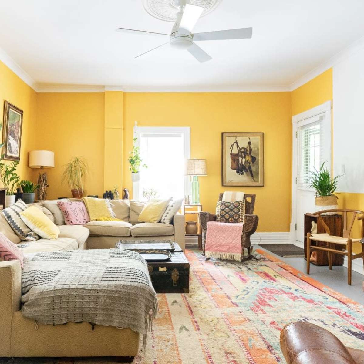A cozy living room features a yellow wall, a large sectional sofa with various textured throws, an acoustic guitar, and a vintage rug.
