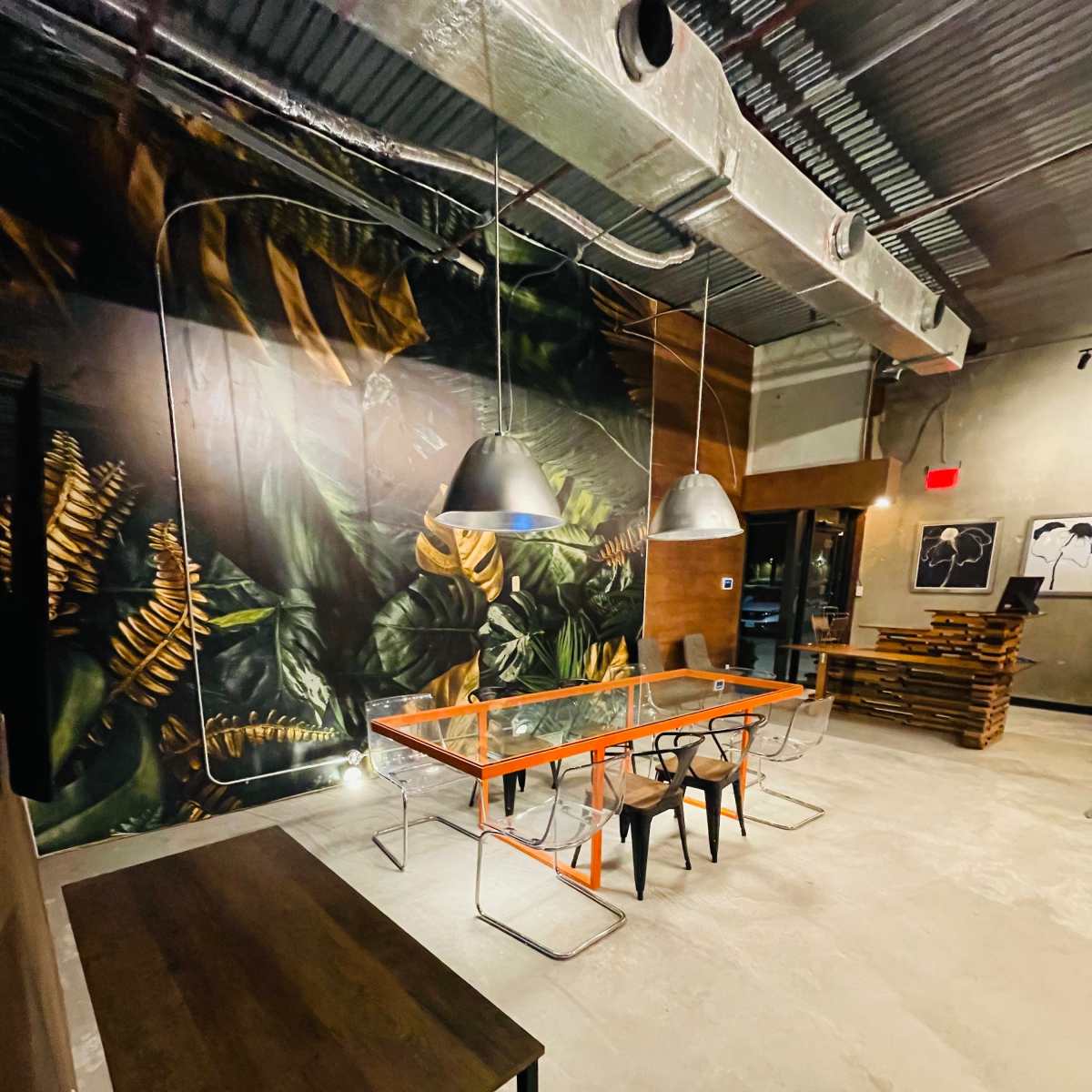 The scene shows a modern interior space featuring a large mural of plants on one wall, a long orange table surrounded by black chairs, and a wooden counter in the background.