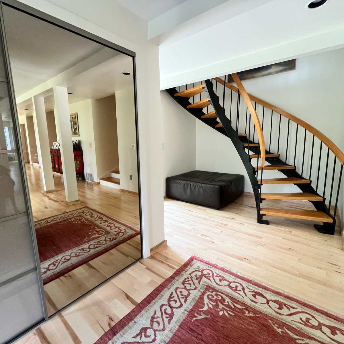 A modern hallway featuring a spiral staircase, a large mirror, and a rug on a wooden floor.
