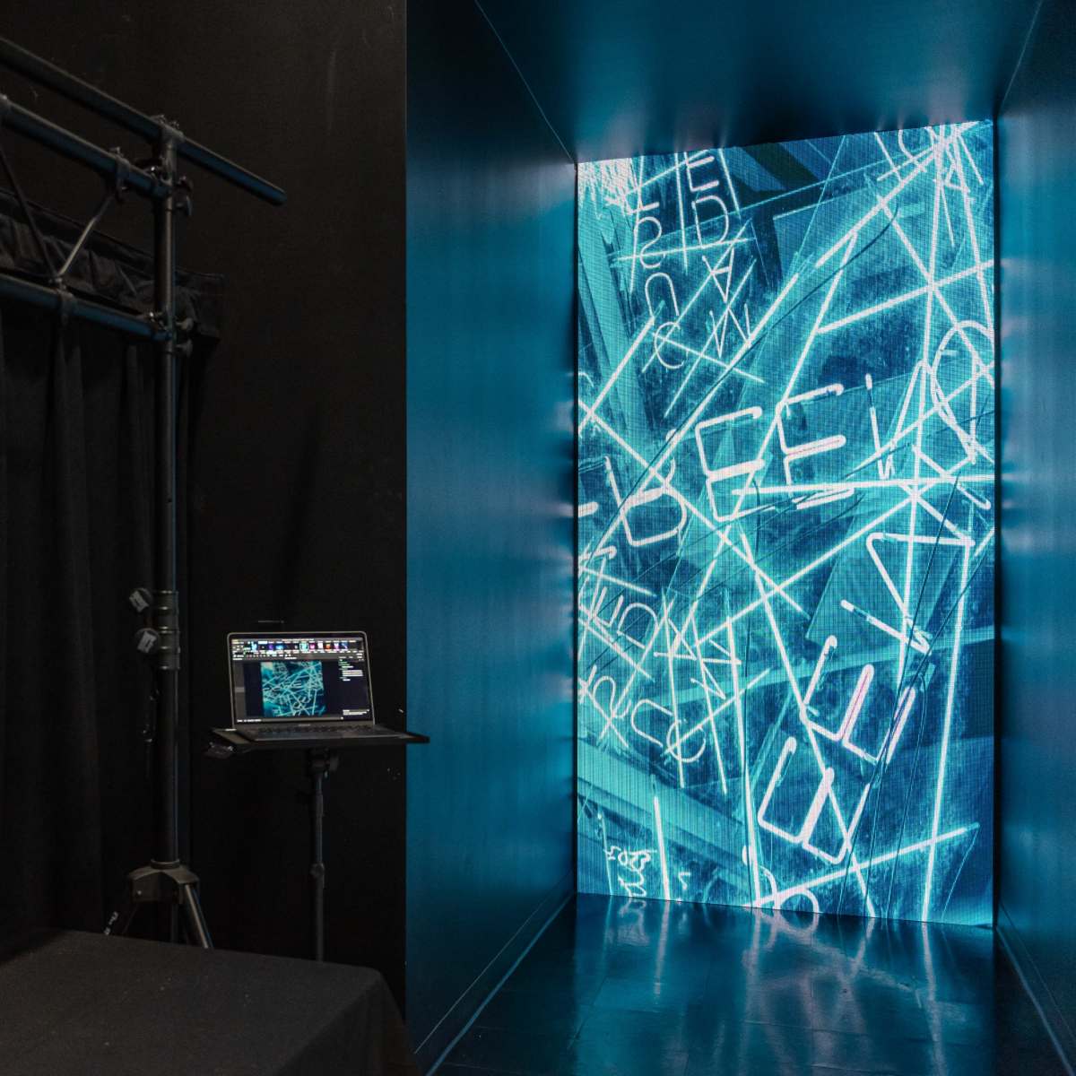 A darkened corridor leading to a brightly lit wall displaying blue digital text, with a black curtain and a laptop on a stand nearby.