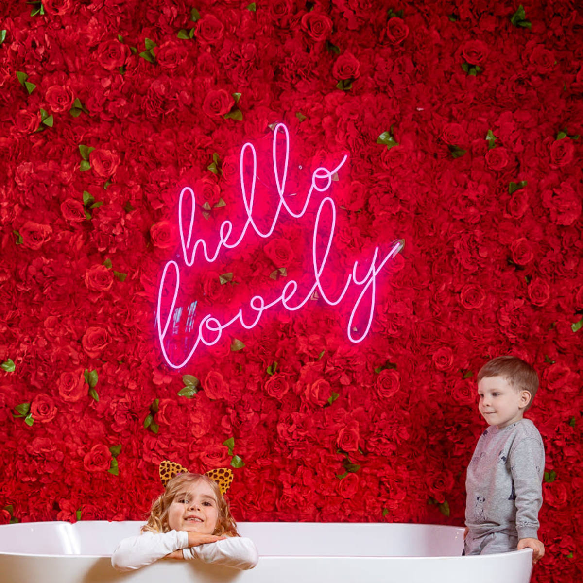 Two children are playing in a white bathtub in front of a vibrant red floral backdrop with a neon sign that says "hello lovely."