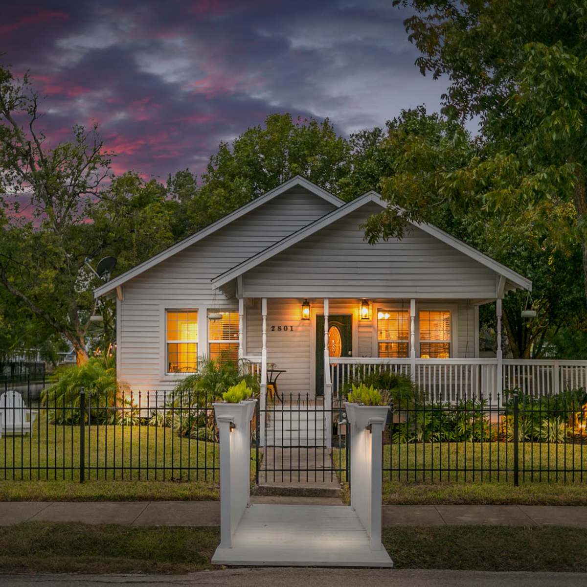 A single-story gray house with a porch is surrounded by greenery and a white picket fence under a dramatic sky at dusk.