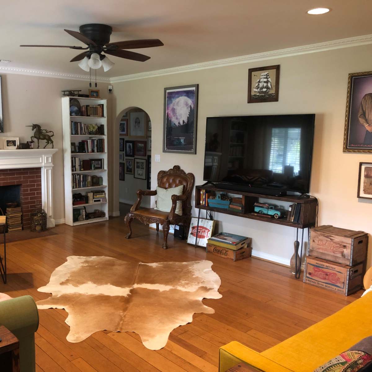 A living room features a fireplace, a television, a cowhide rug on wooden flooring, and a mix of furniture including a green sofa and a vintage armchair.