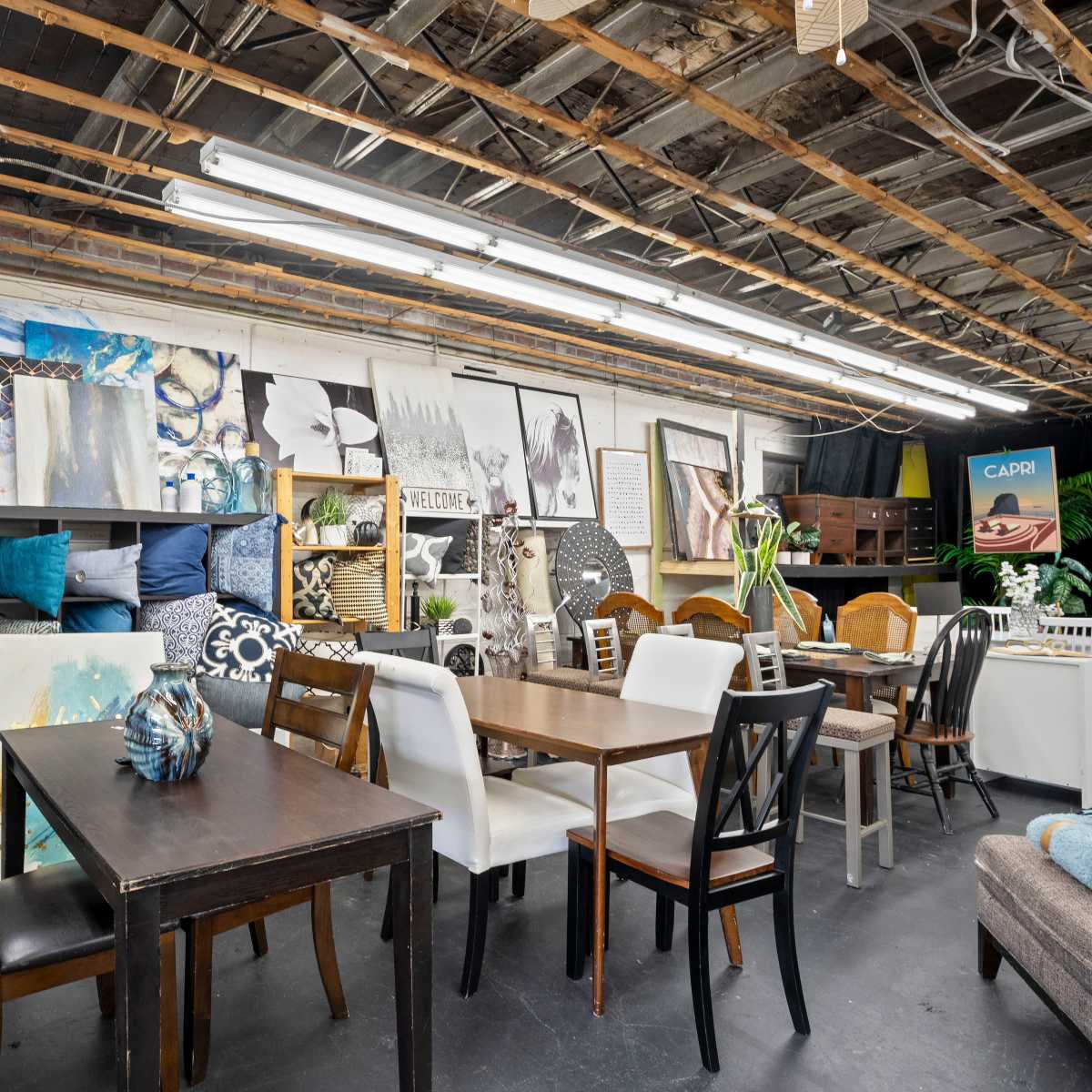 A well-organized interior space displaying various furniture pieces, artwork, and decorative items against a backdrop of exposed beams and fluorescent lighting.