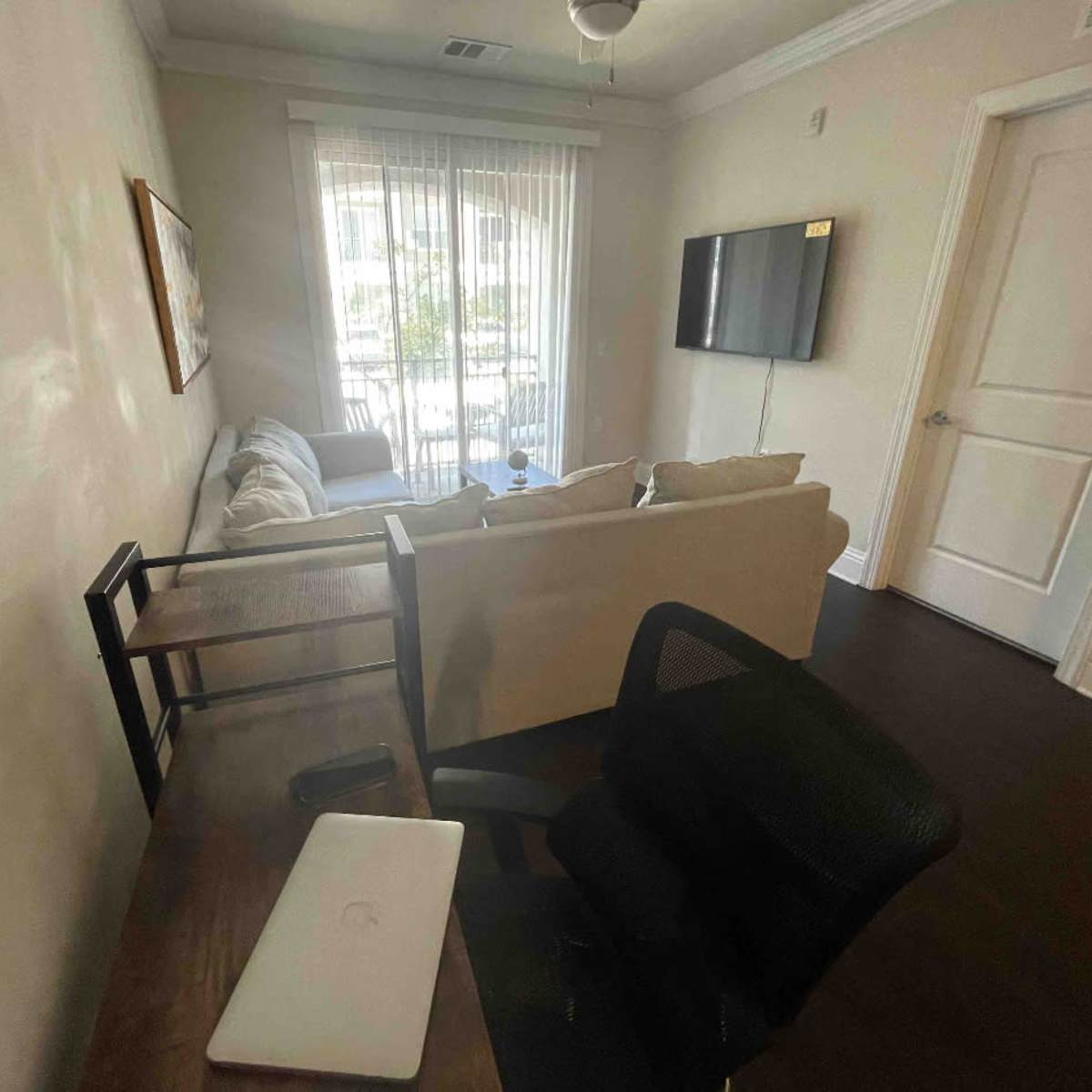 A small living area features a couch, a desk with a laptop, and a television mounted on the wall, with natural light streaming through the window.
