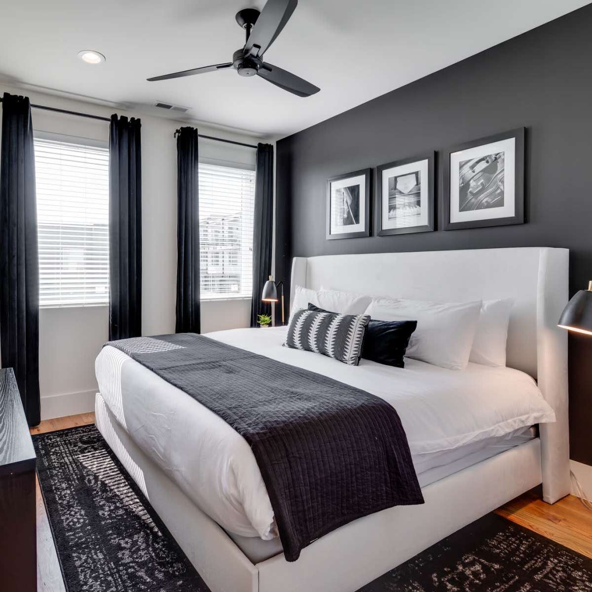 A modern bedroom features a white bed with a dark throw blanket, a television mounted on the wall, and large windows dressed with black curtains.