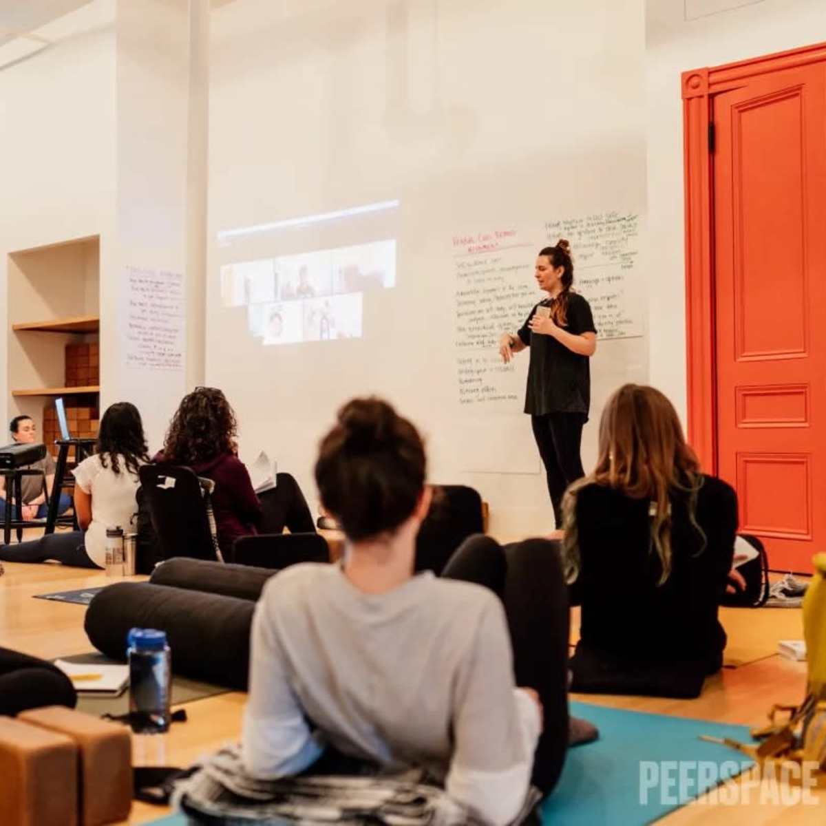 A group of participants sits on yoga mats in a studio while a speaker presents in front of them with a projected screen.