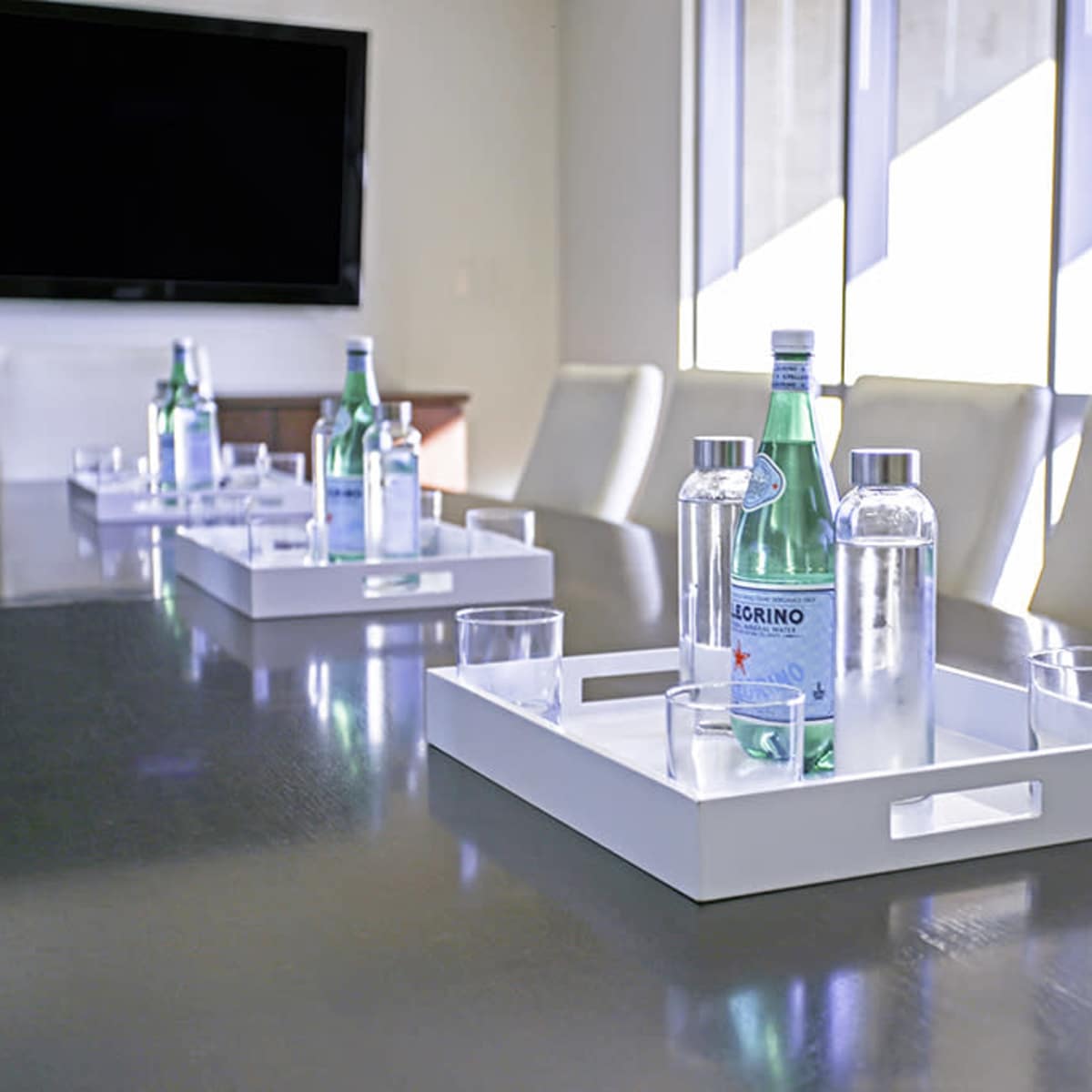 A conference table is set with trays containing bottles of sparkling water and glasses, with large windows in the background.