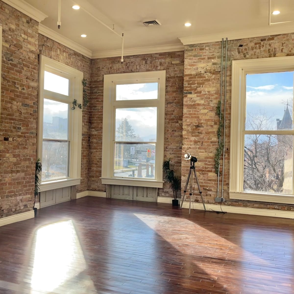 A spacious, well-lit room with exposed brick walls, large windows, and polished wooden floors.