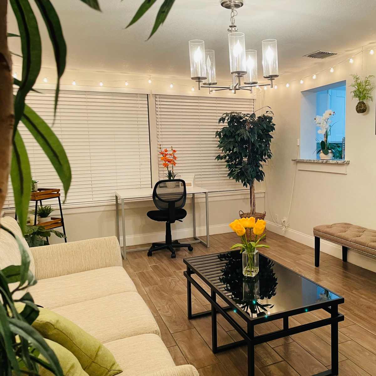 A well-lit living room features a light-colored sofa, a glass coffee table with a vase of orange flowers, and a decorative plant, with a desk and chair in the background.