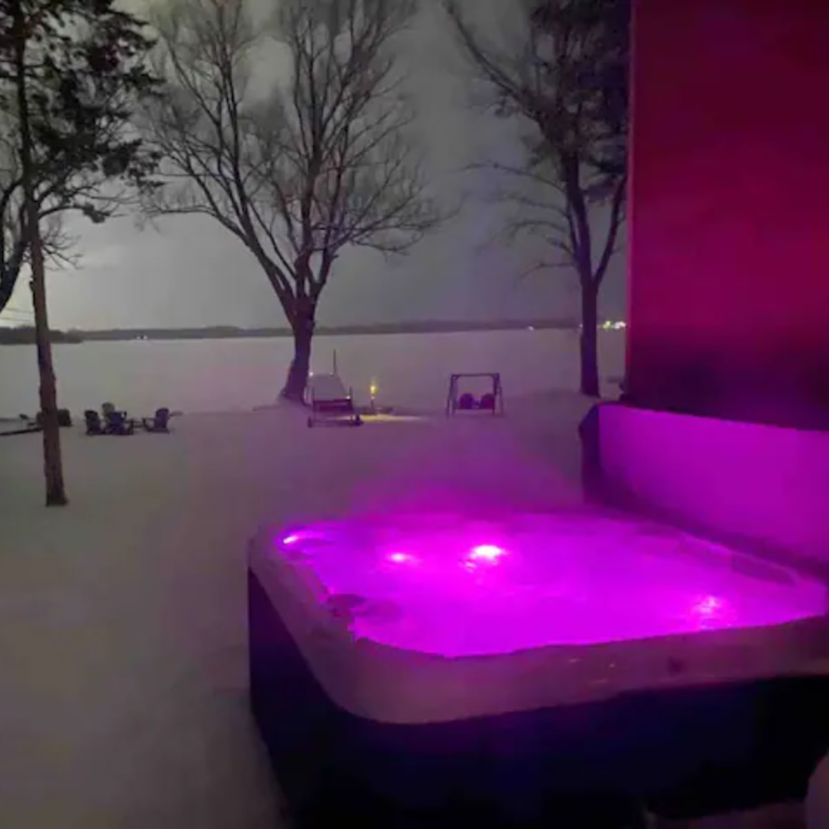 A hot tub with purple lights sits in the snowy yard near a lake, framed by leafless trees.